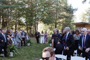 Outdoor wedding ceremony in a wooded setting with guests seated on chairs on a grassy lawn.