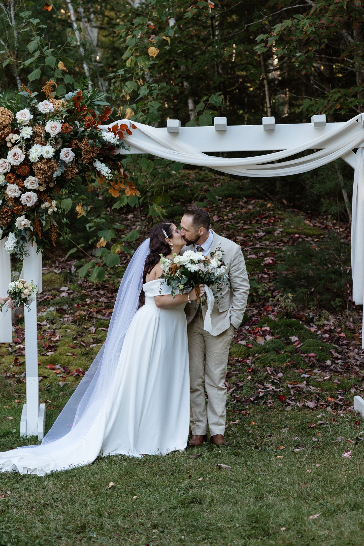 Outdoor wedding ceremony under a white draped arch with floral arrangements in a garden setting.