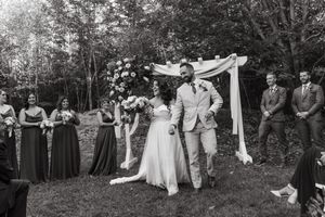 A black and white outdoor wedding ceremony under a draped fabric arch with wedding party members standing in a garden setting.