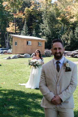 A wedding couple stands on a green lawn with a rustic wooden cabin and autumn trees in the background.