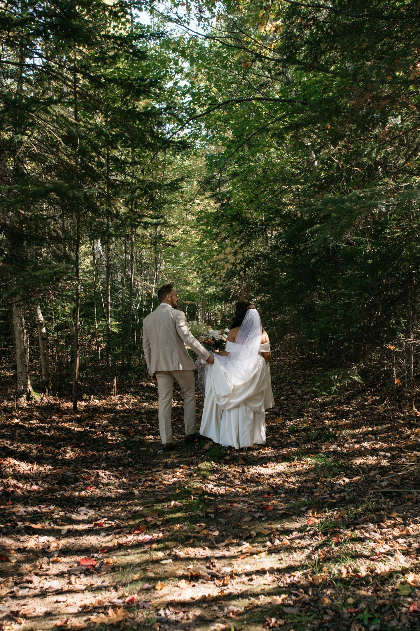 A couple walking through a sunlit forest path with fallen autumn leaves during a wedding ceremony.