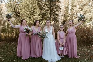 A bridal party in dusty rose pink dresses and a white wedding gown pose together in a wooded outdoor setting.