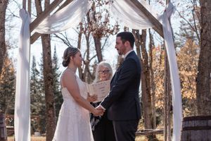 A wedding ceremony takes place under a white draped arbor in an autumn outdoor setting with trees in the background.