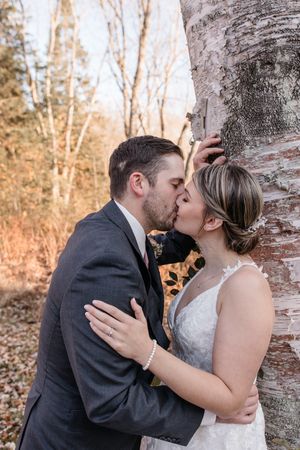 A romantic wedding couple shares tender kisses against a birch tree trunk in a winter forest setting.