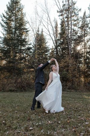 A couple in wedding attire dances playfully in a field surrounded by evergreen trees at sunset.