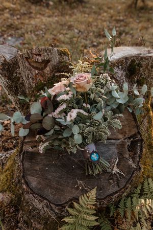 A rustic wedding bouquet with pink roses and eucalyptus leaves rests on a weathered wooden log in a forest setting.