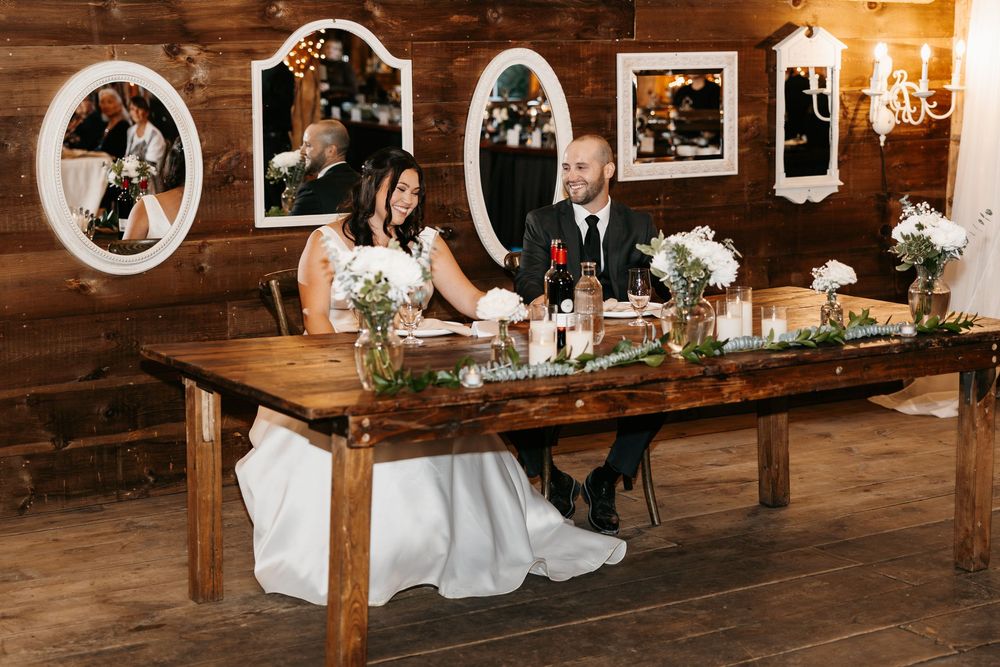 A couple sits at a rustic wooden farm table decorated with flowers inside a barn with family photos on wood paneled walls.