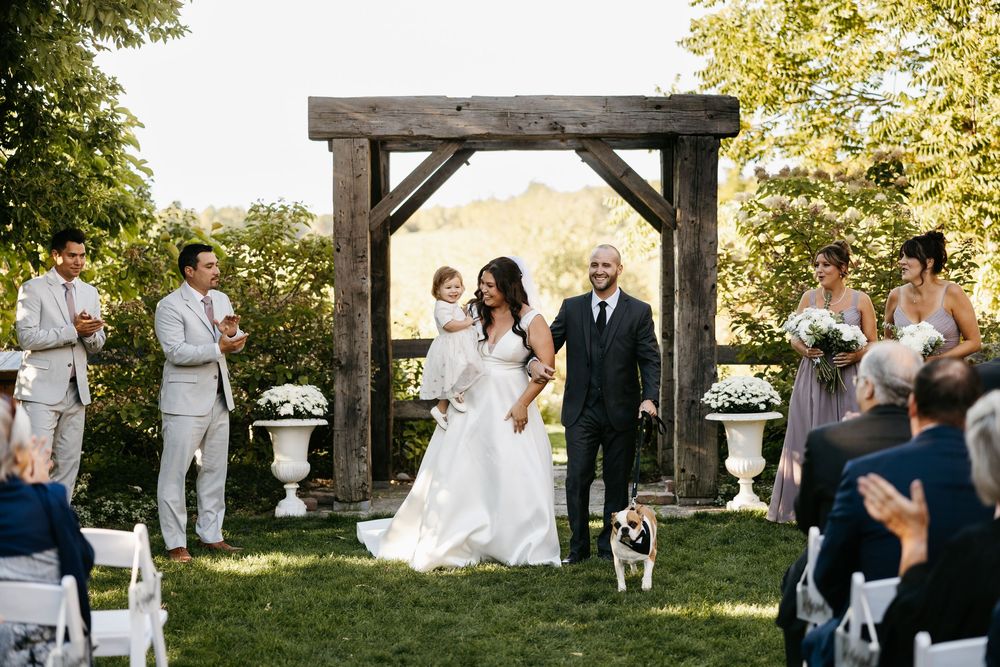 A rustic outdoor wedding ceremony unfolds beneath a wooden pergola surrounded by lush greenery and seated guests.