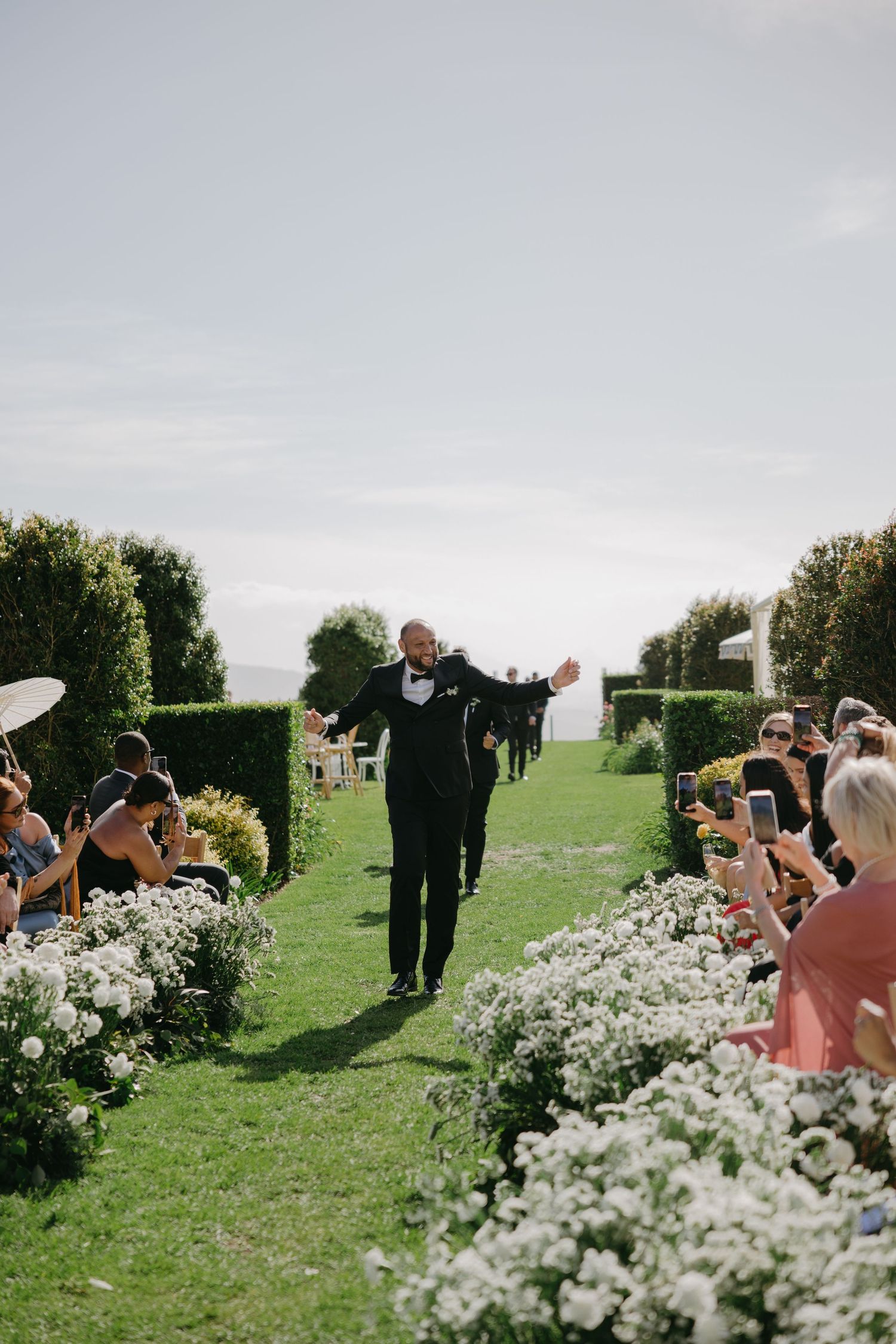 Wedding guests take photos along a garden path lined with blooming white flowers on a green hillside.
