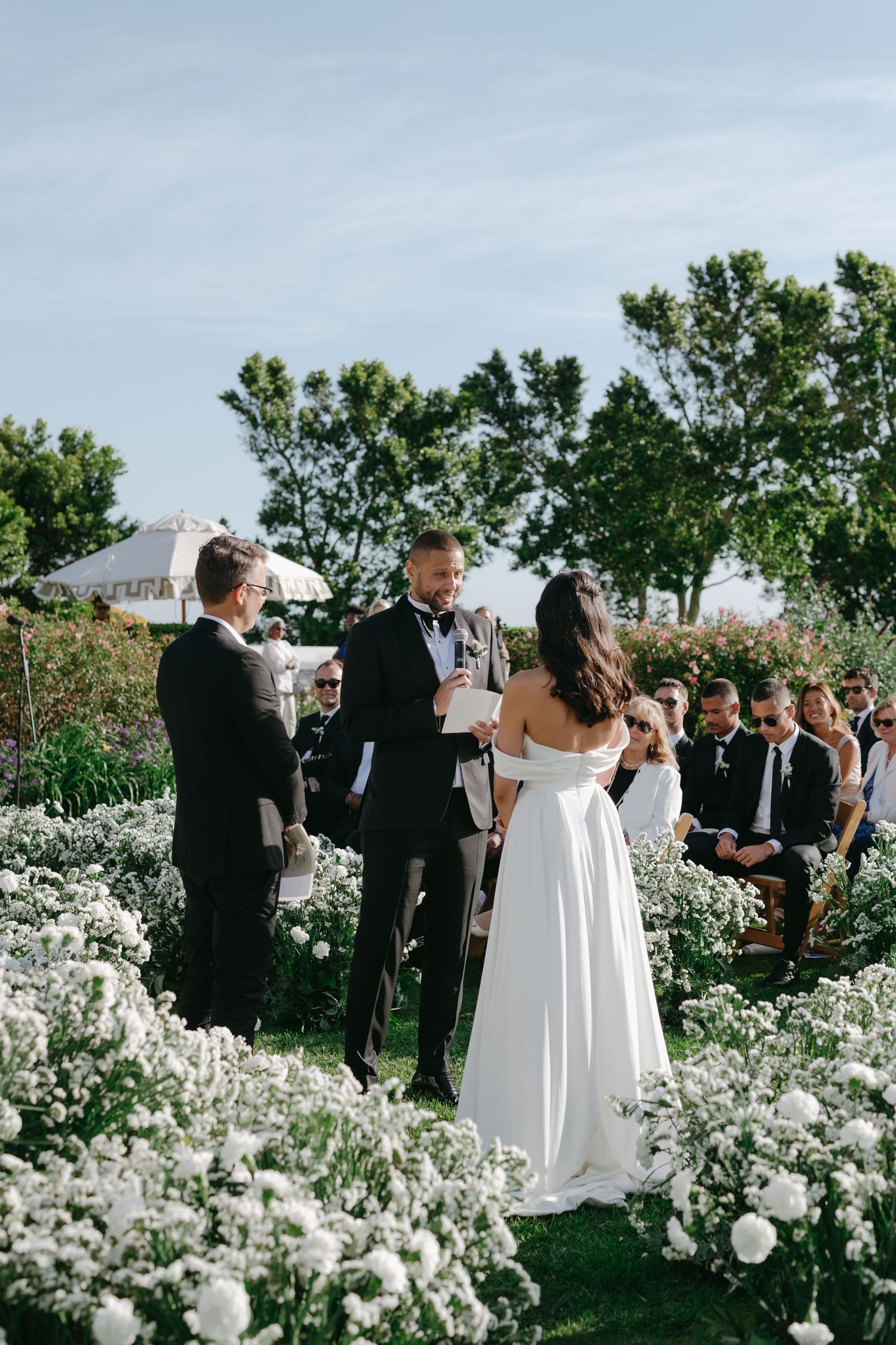 Elegant outdoor wedding ceremony surrounded by lush white floral arrangements and greenery in a garden setting.