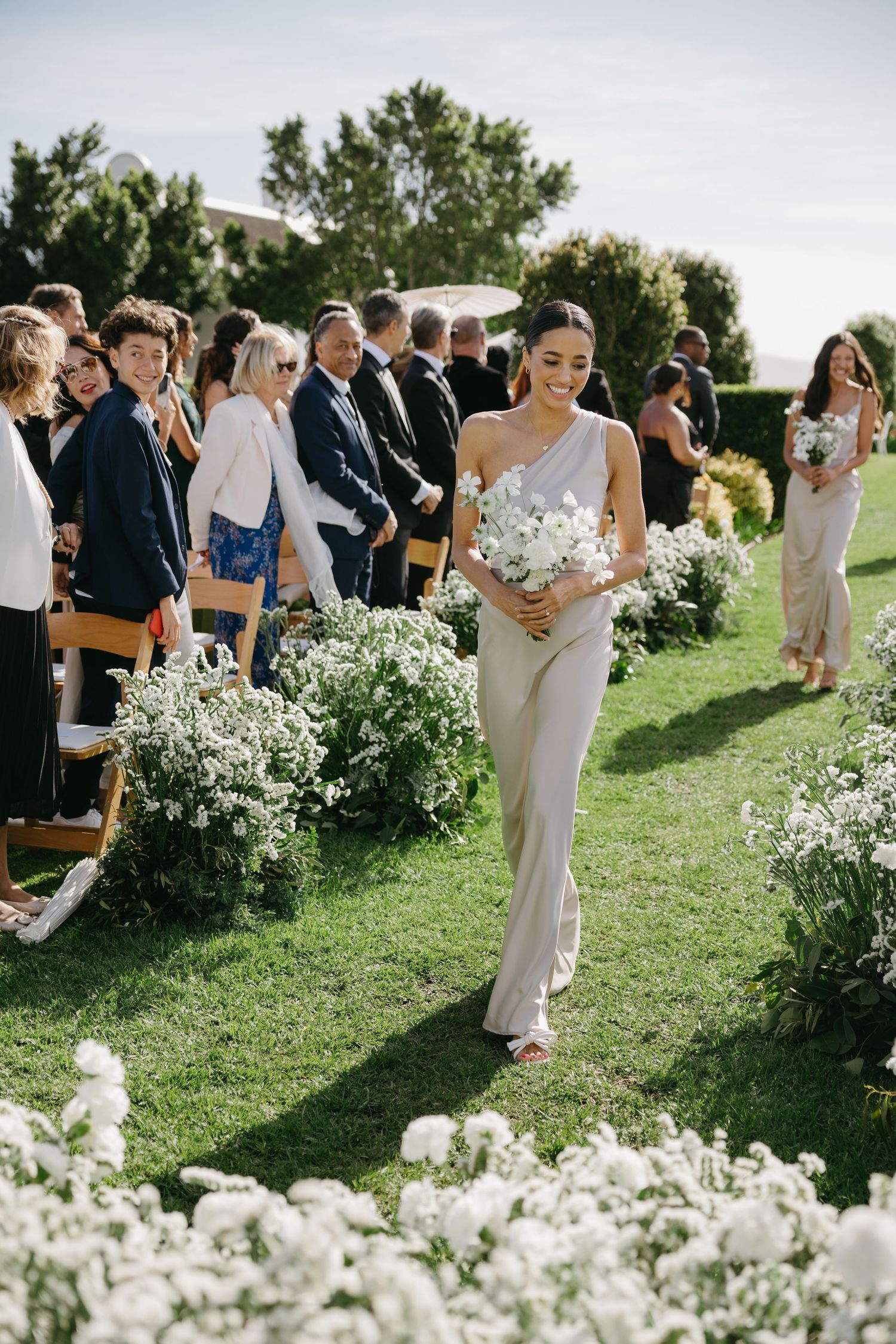 Wedding guests gather on a lush green lawn surrounded by delicate white flowers during an outdoor summer celebration.