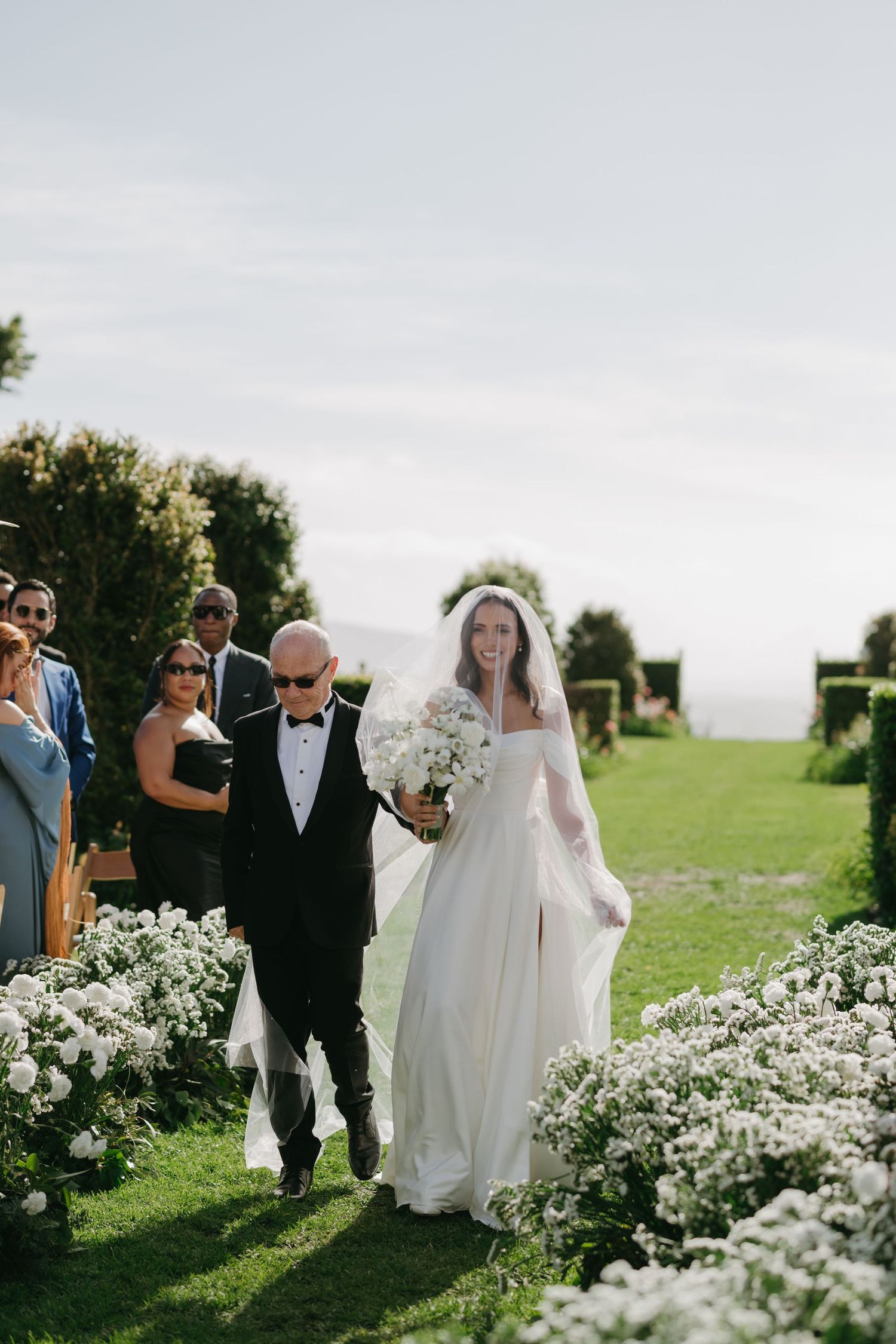 Wedding ceremony procession along a garden path lined with beautiful white flowers on a sunny summer day.