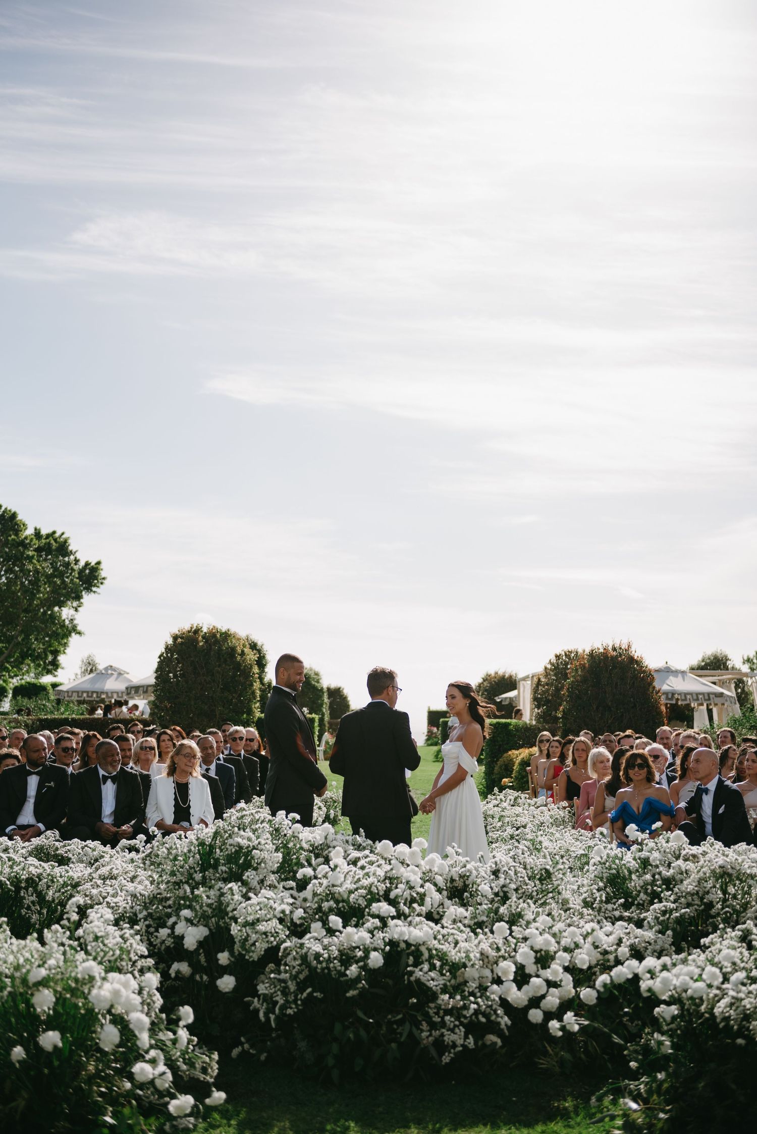 Guests gathered outdoors at an elegant garden wedding ceremony with white floral arrangements and natural greenery.
