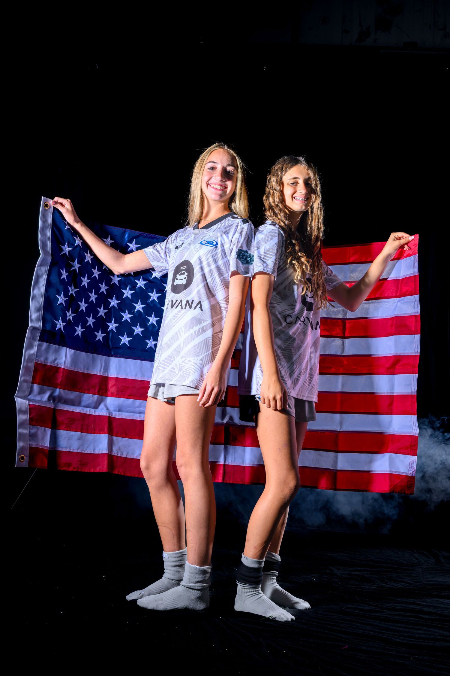 Two people in white t-shirts pose playfully in front of an American flag backdrop.
