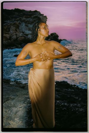 Artistic sunset photoshoot at rocky beach with figure draped in flowing beige fabric against dramatic pink and purple sky.