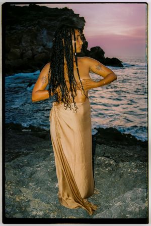 A person in a flowing gold dress stands on rocky coastal cliffs at sunset with waves crashing against the shore.
