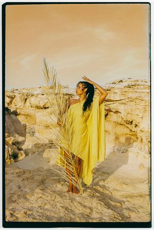 A figure in a flowing yellow dress poses with palm fronds against sandy cliffs during golden hour.