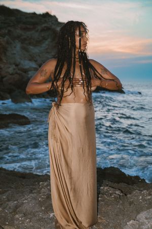 A silhouette stands on rocky coastline at sunset wearing a draped dress with waves crashing in the background.
