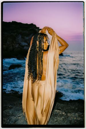 A figure draped in golden fabric stands against a rocky coastline at sunset with crashing waves in the background.