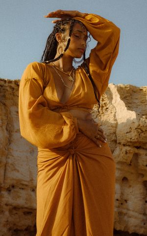 A figure in a flowing mustard yellow dress poses dramatically against a rocky cliff backdrop in warm sunlight.
