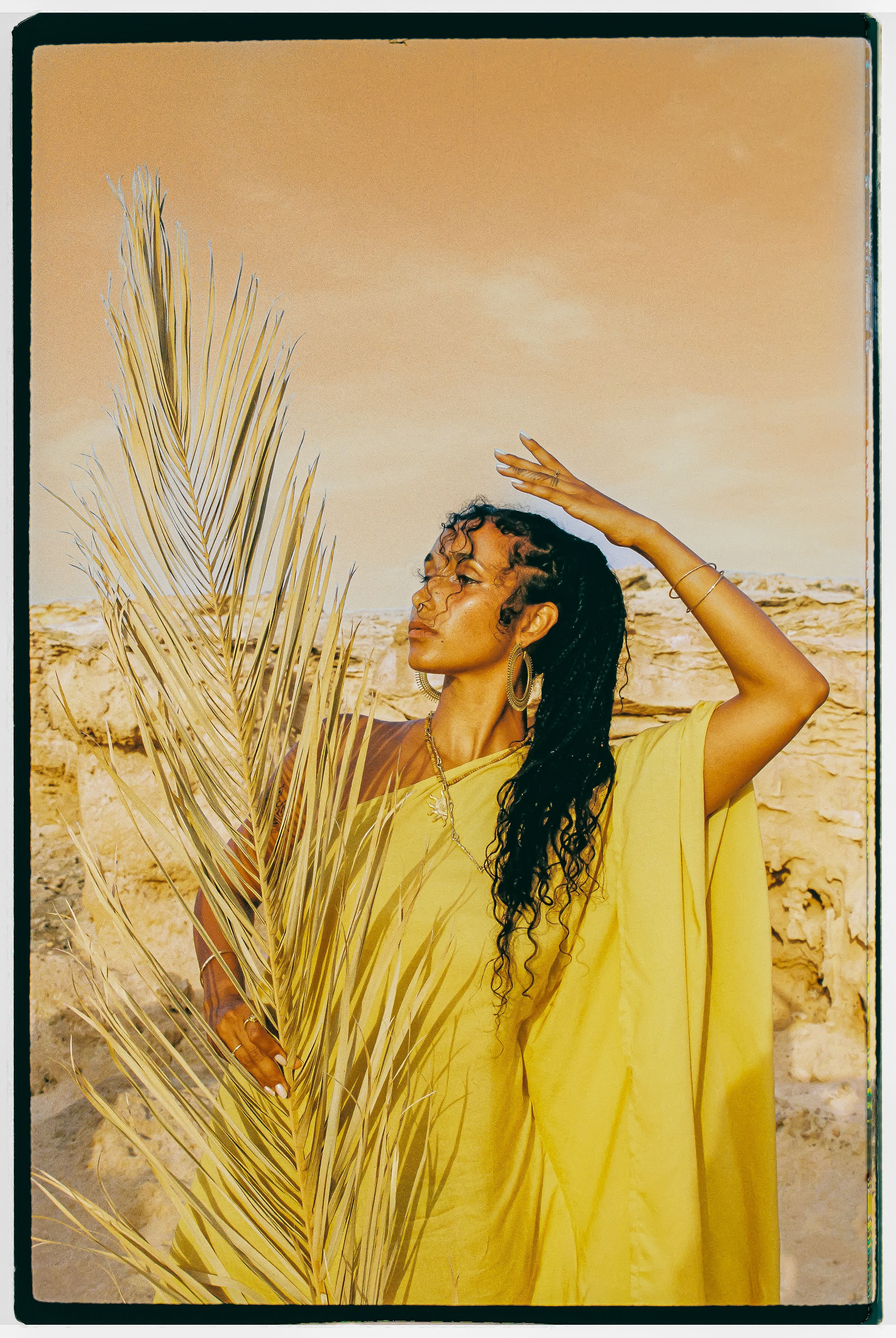 Figure in bright yellow dress poses expressively among dried palm fronds.