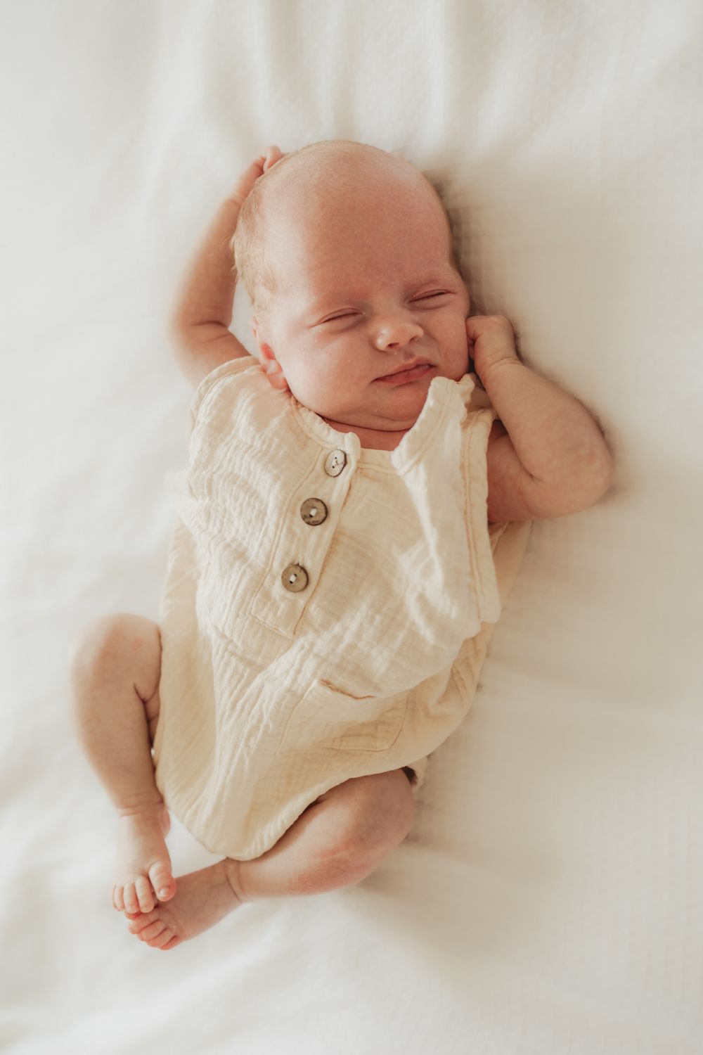 A peaceful sleeping newborn wearing a cream-colored outfit lies on white bedding.
