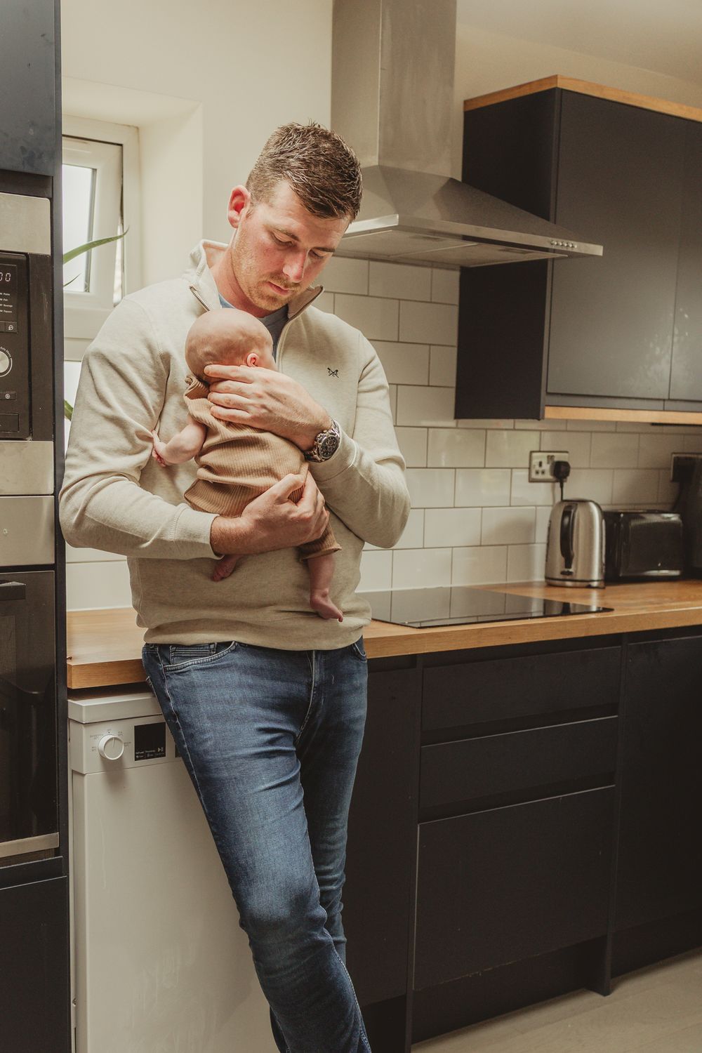 A parent in casual attire holds a baby while standing in a modern kitchen with subway tile.