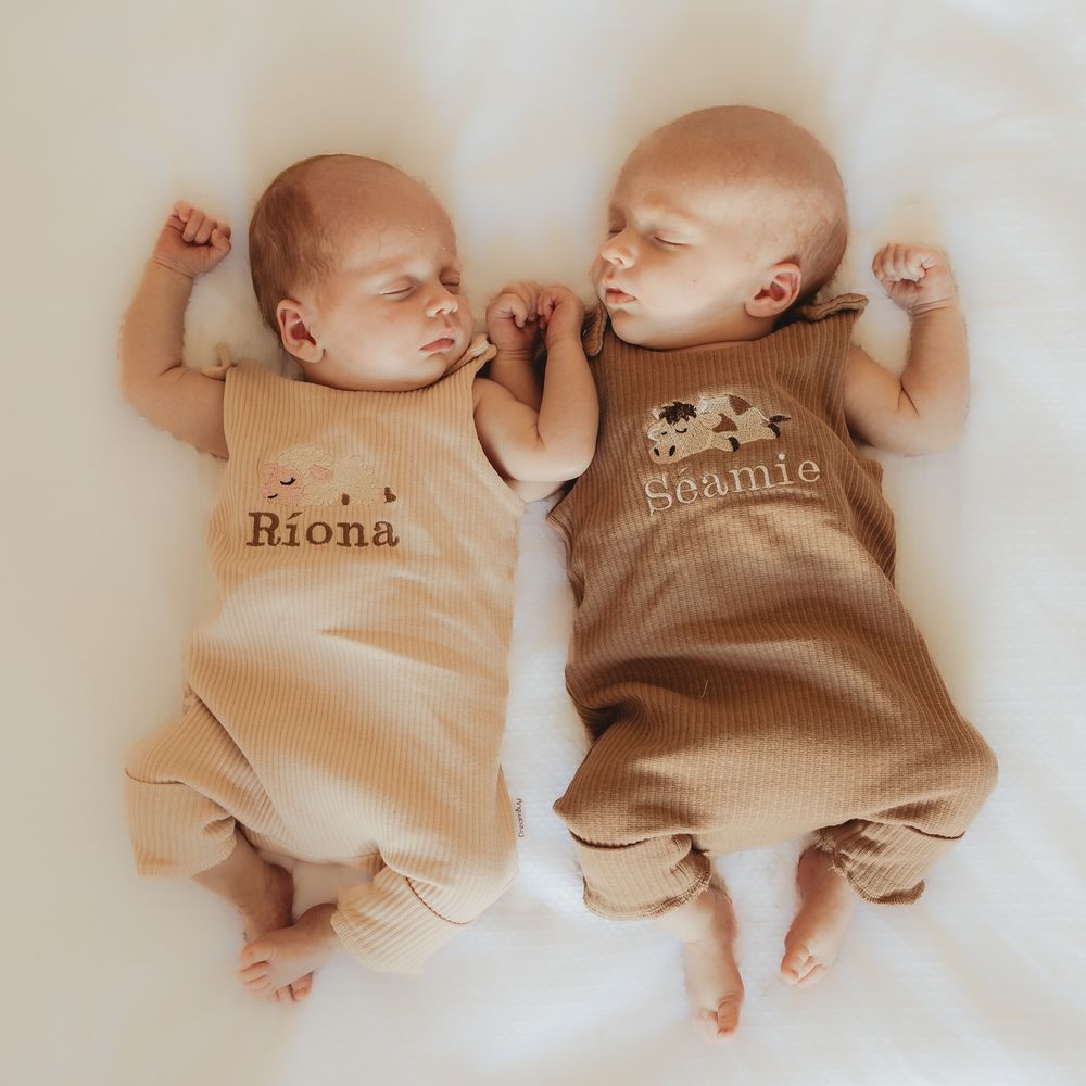 Two sleeping newborn babies in matching onesies lying side by side on white bedding.