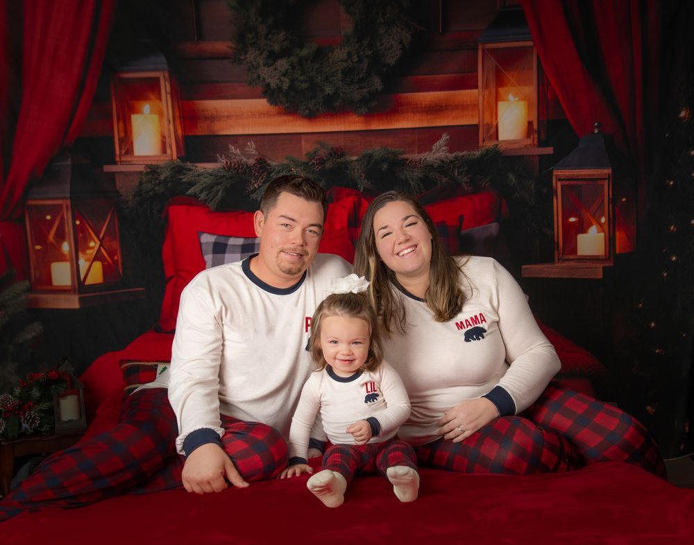 A family in matching white shirts and plaid pajamas poses for holiday photos in front of a red backdrop with lanterns.