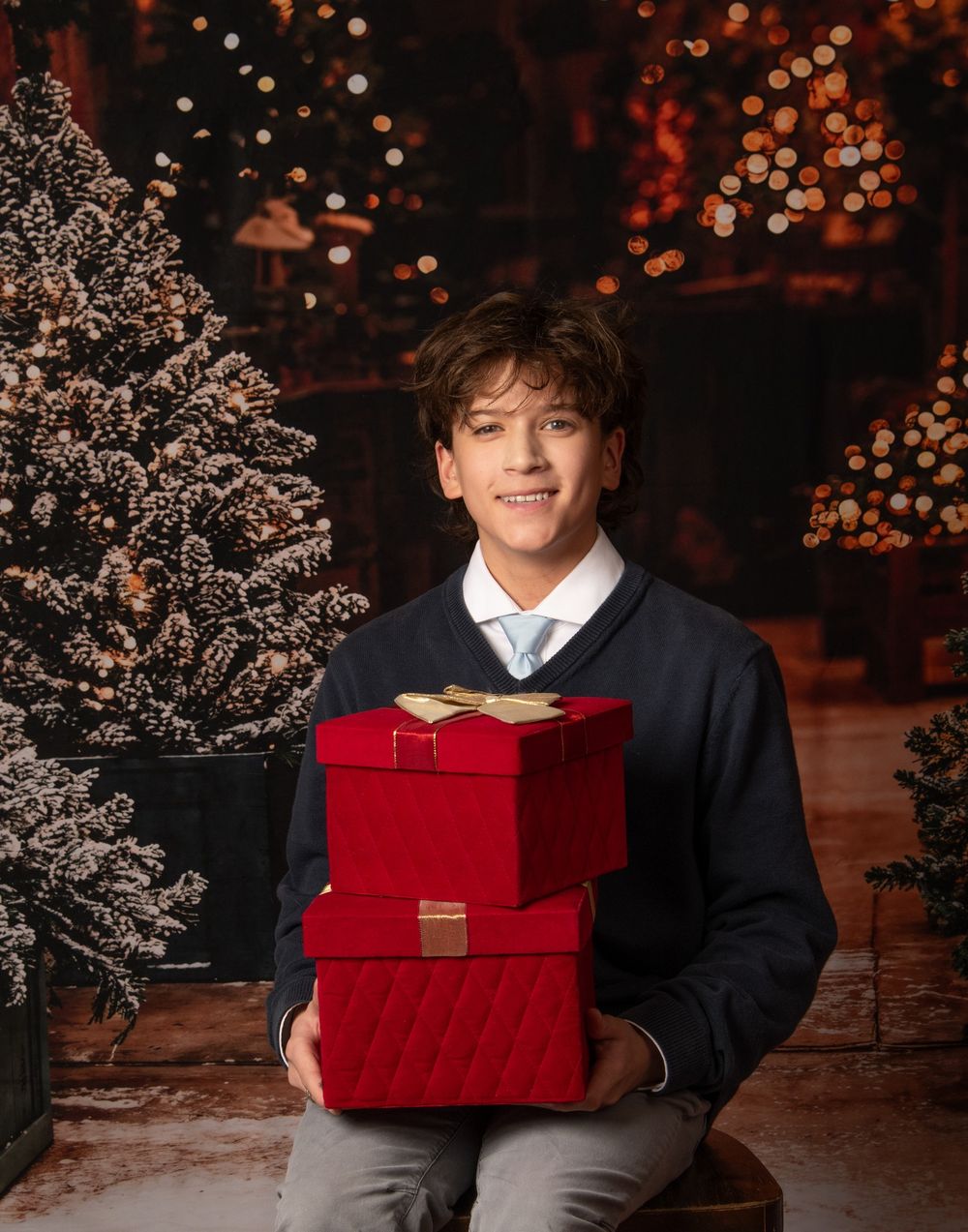 A festive holiday scene with someone holding a bright red gift box against a backdrop of twinkling lights and evergreen branches.
