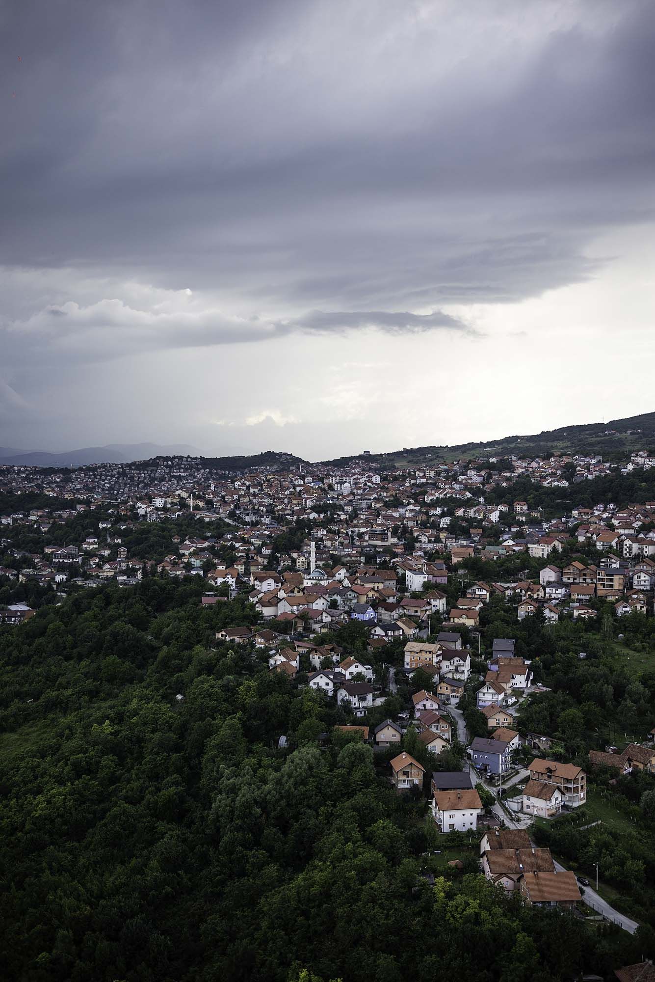 Aerial view of a hillside town with white houses nestled among green forests under dramatic stormy skies.