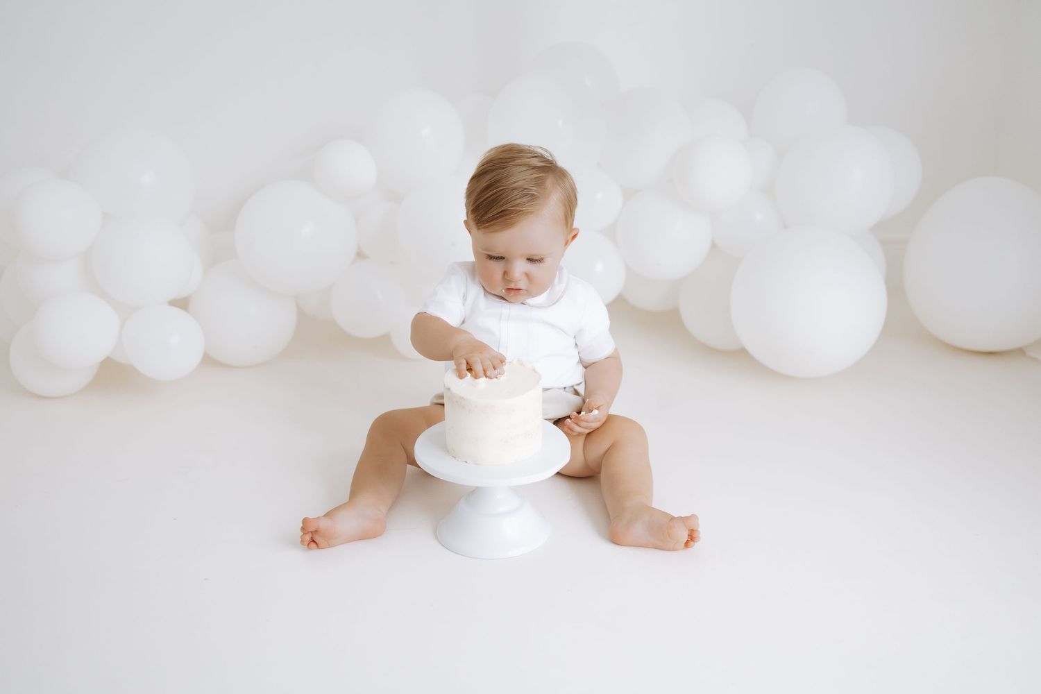 A small child in white sits with a birthday cake against a backdrop of white balloons in a bright studio setting.