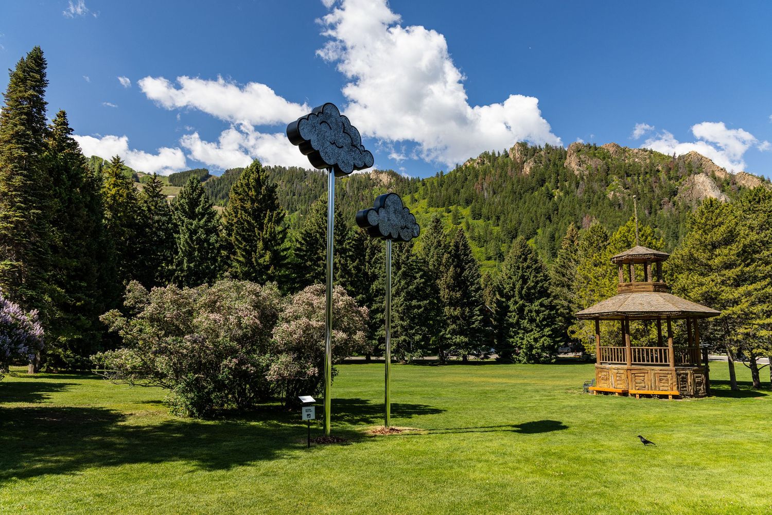 A panoramic mountain landscape with lush green meadows and a wooden gazebo structure under bright blue summer skies.