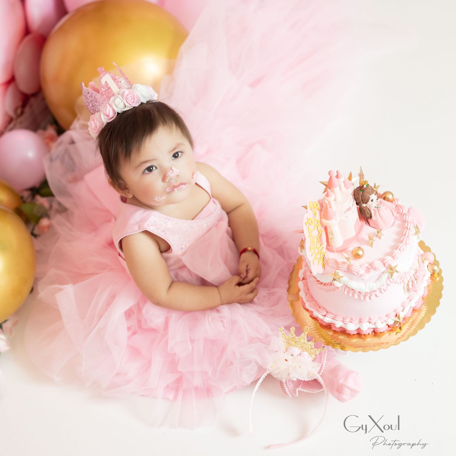 Baby in pink tulle dress sits on floor with birthday cake during first birthday cake smash photo session.