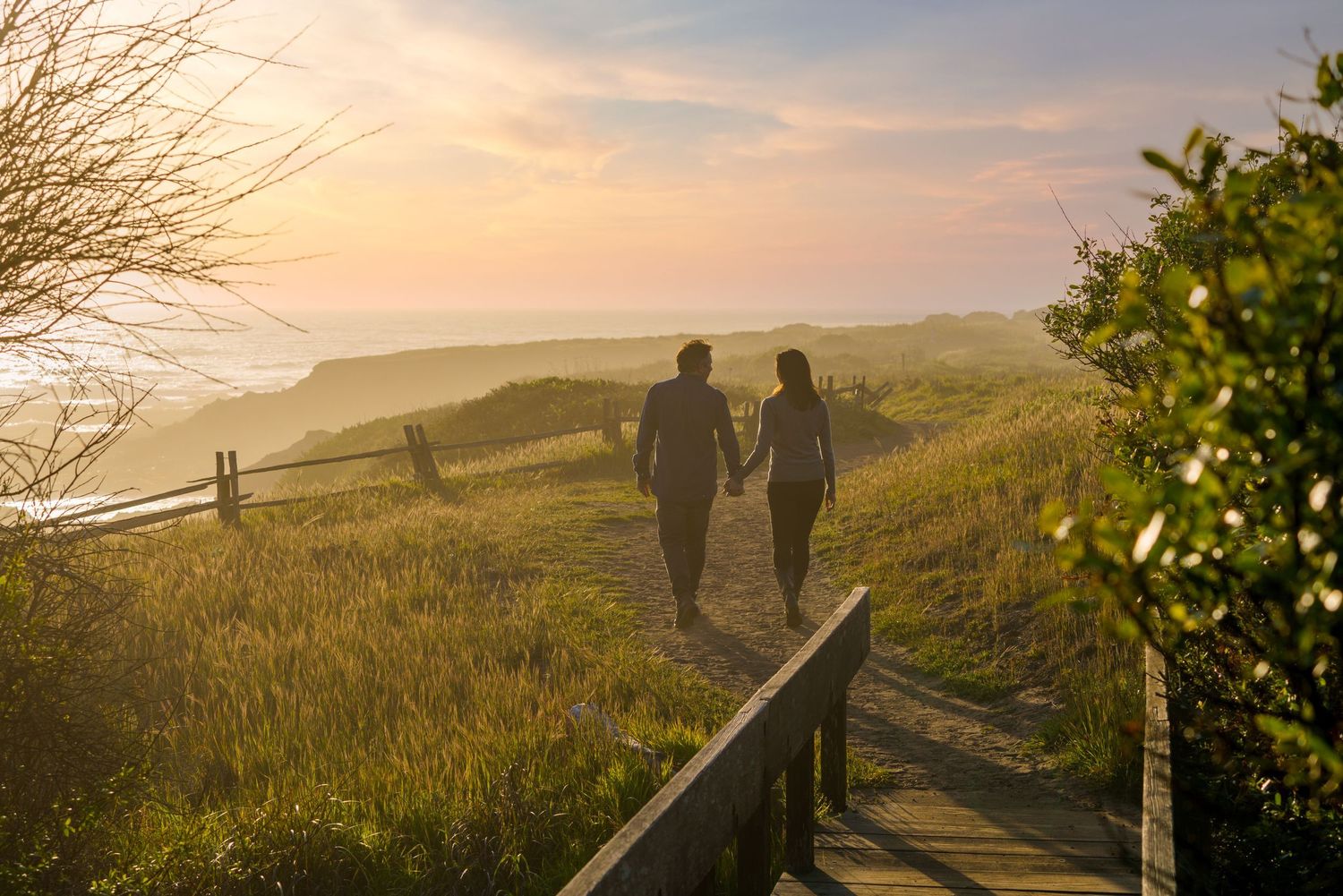 Silhouetted figures walk along a scenic hilltop trail during golden hour as the sun sets over rolling hills.