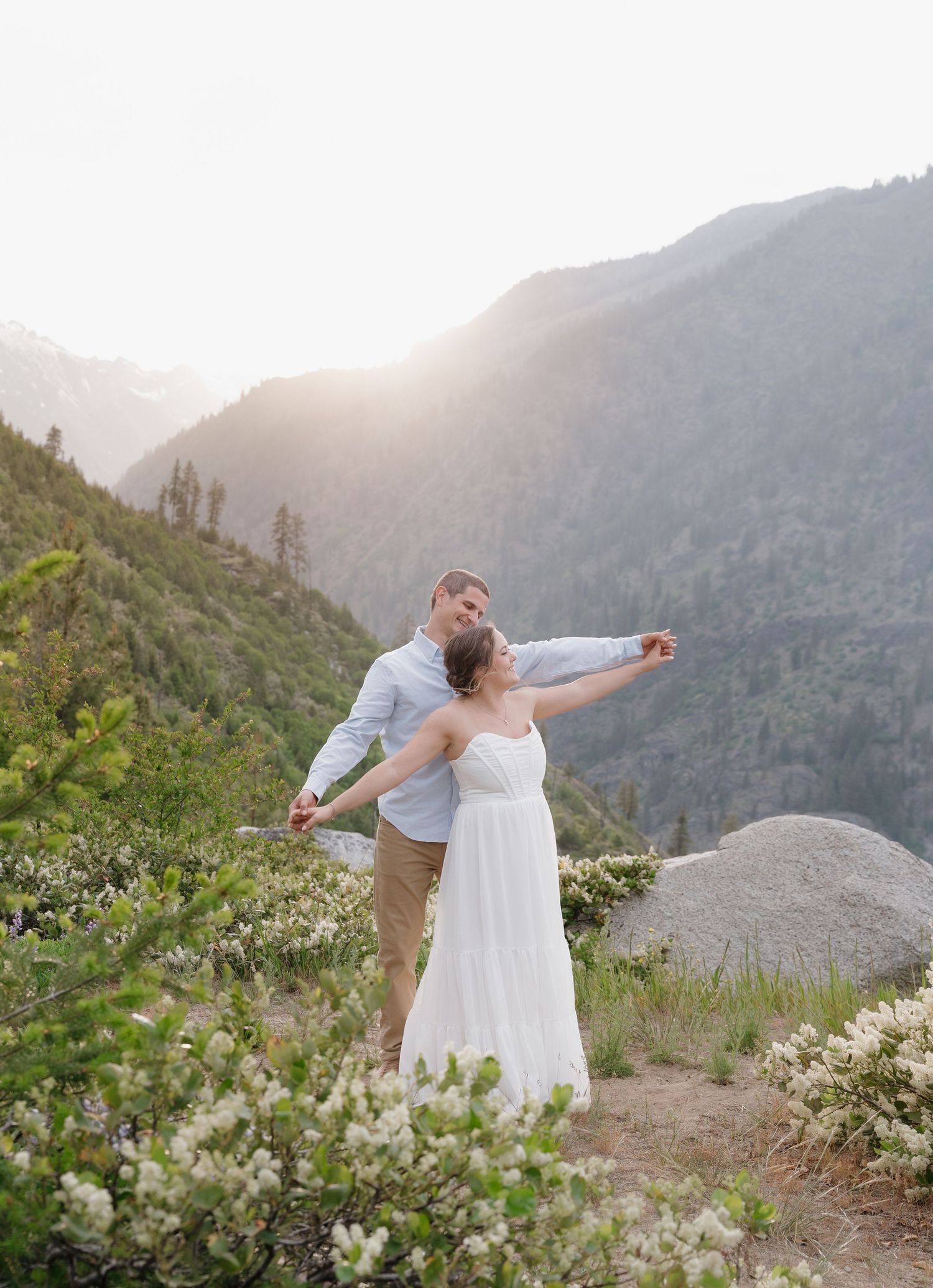 A couple embraces on a mountain overlook at sunset with wildflowers in the foreground and misty peaks in the distance.