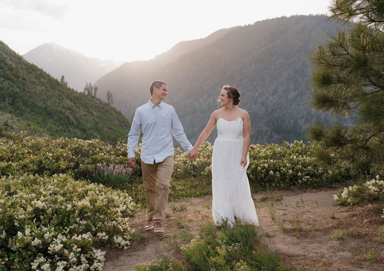 Couple walking along a mountain trail surrounded by wildflowers at sunset in a scenic natural landscape.