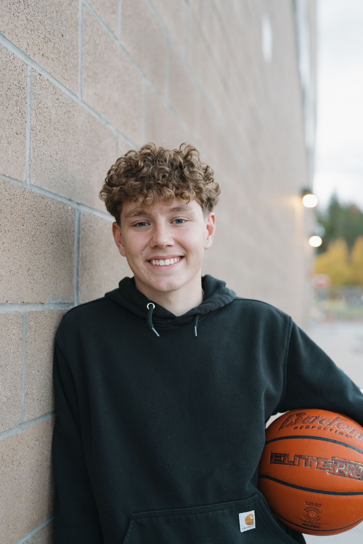 A person in a black hoodie leans against a wall while holding a basketball.