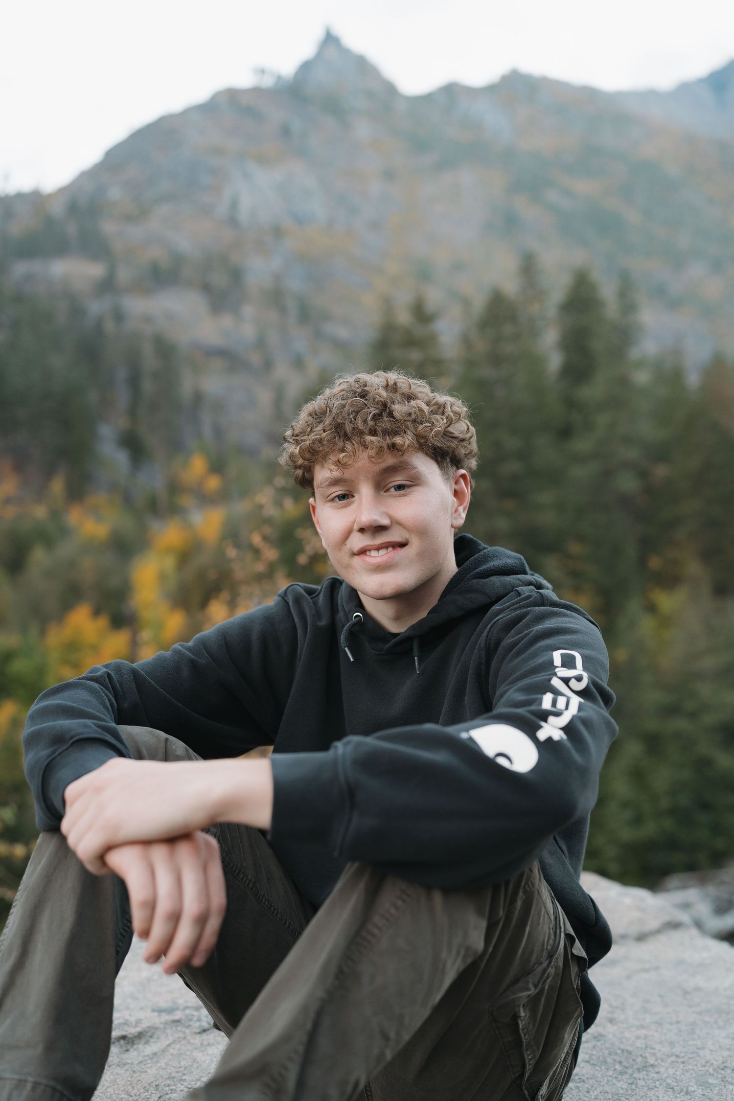 Person in black hoodie sitting on rocky outcrop with mountainous backdrop.