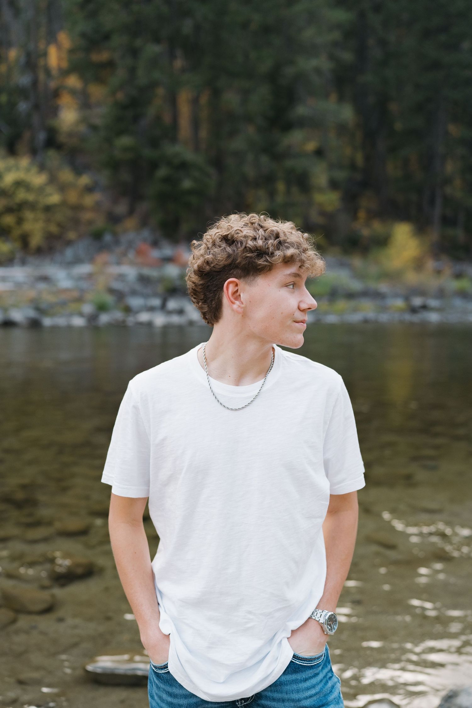 Person in white t-shirt standing by riverbank with autumn forest in background.