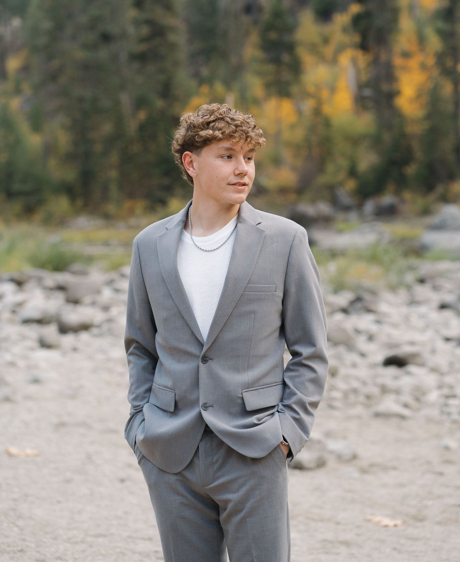A person in a gray suit poses outdoors against an autumn mountain landscape.