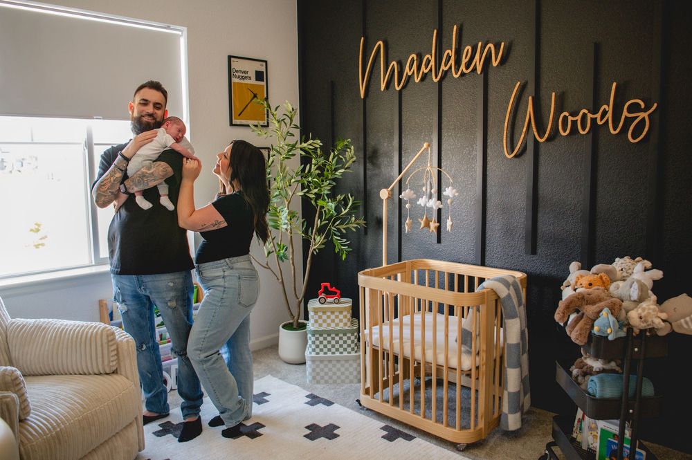 A couple stands together in a cozy nursery room with dark walls and natural wood furniture.