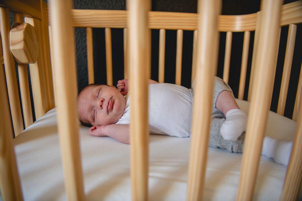 A baby lies in a wooden crib with slats visible, showing a partial view through the crib bars.
