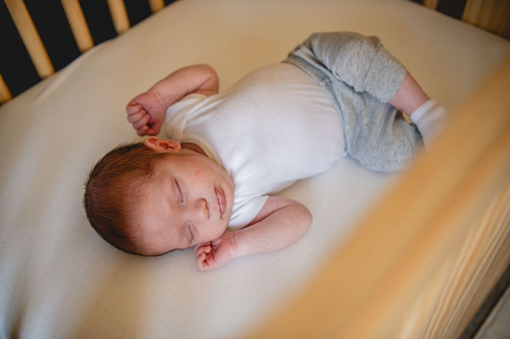 Sleeping baby in white onesie resting peacefully on a crib mattress with white bedding.
