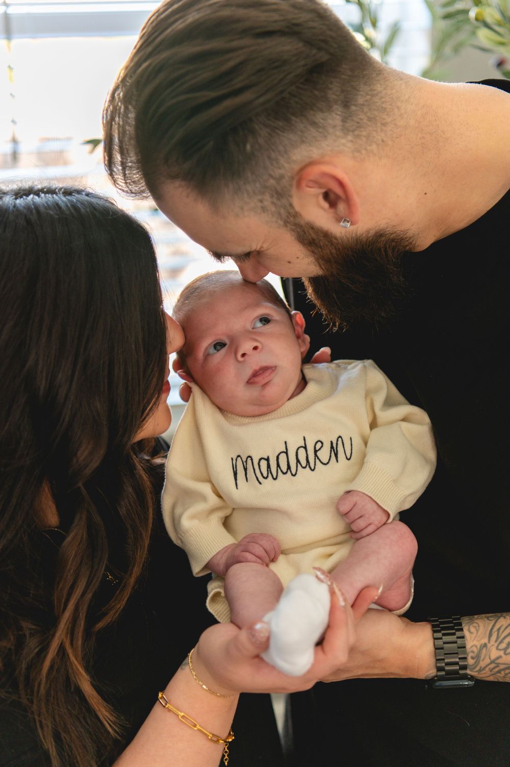 A baby wearing a cream-colored sweater with 'Madden' text is cradled by parents in an intimate family moment.