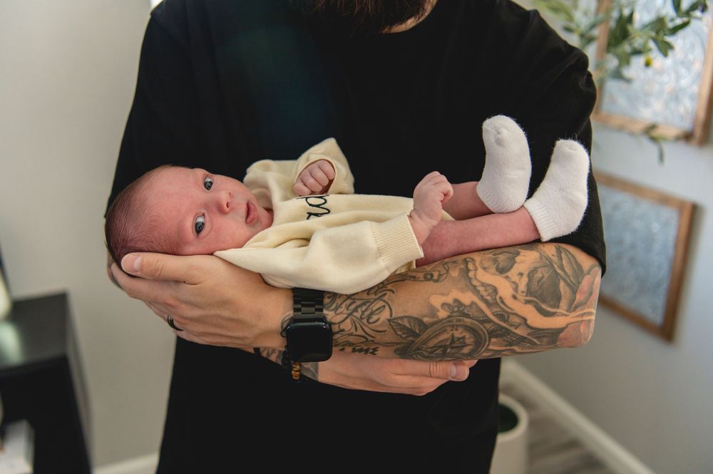 A parent with tattoos gently cradles a sleeping newborn baby in a peaceful home setting.