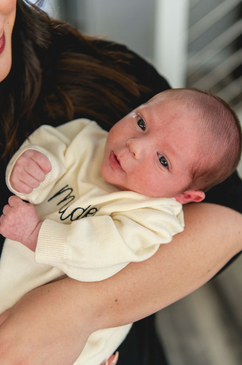 A baby in a cream colored onesie rests peacefully against a caregiver's shoulder.