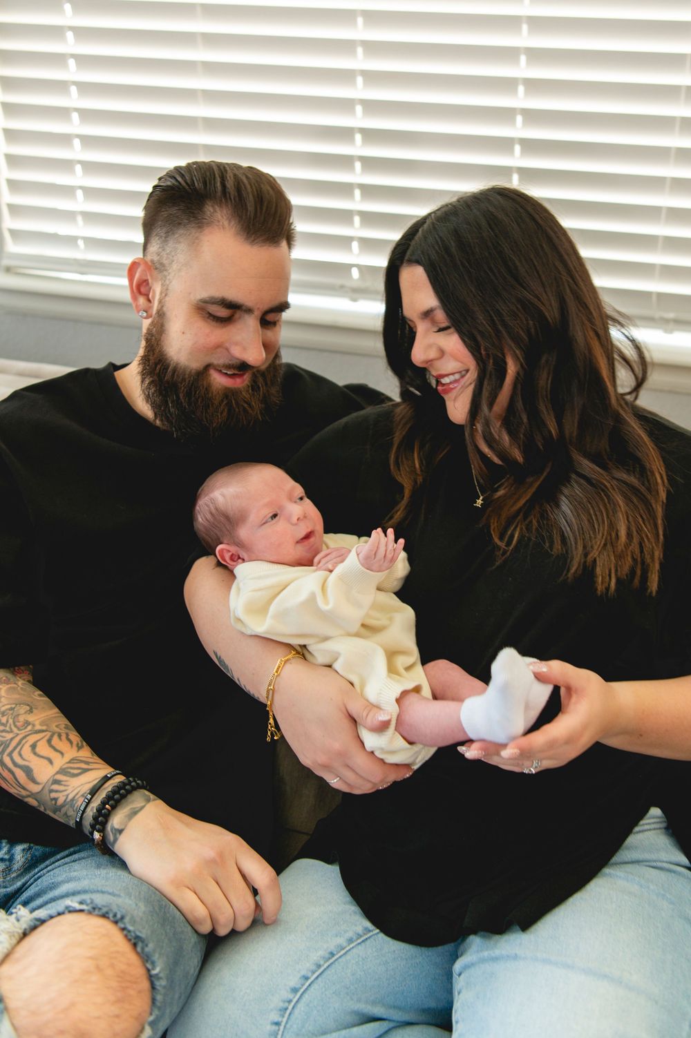 A couple shares a tender moment with their newborn baby while sitting together on a couch by venetian blinds.