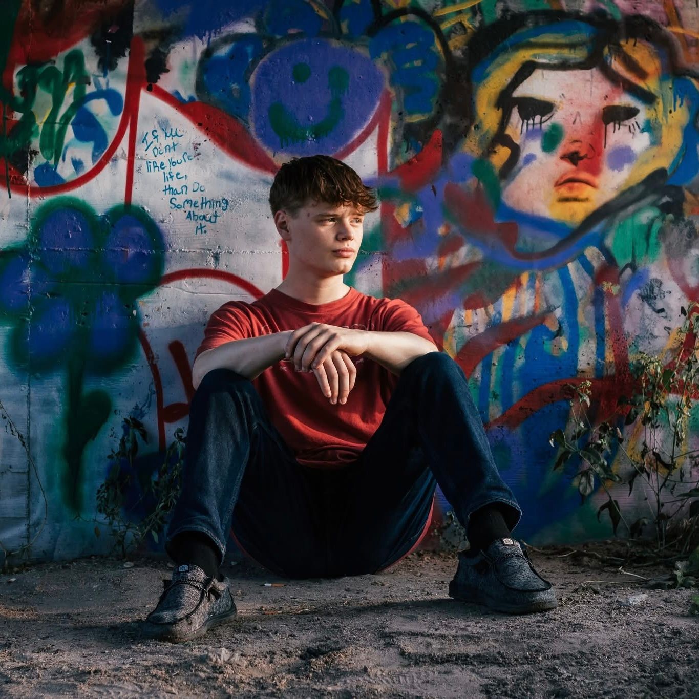 Someone sits against a colorful graffiti wall wearing a red shirt and dark pants in an urban setting.