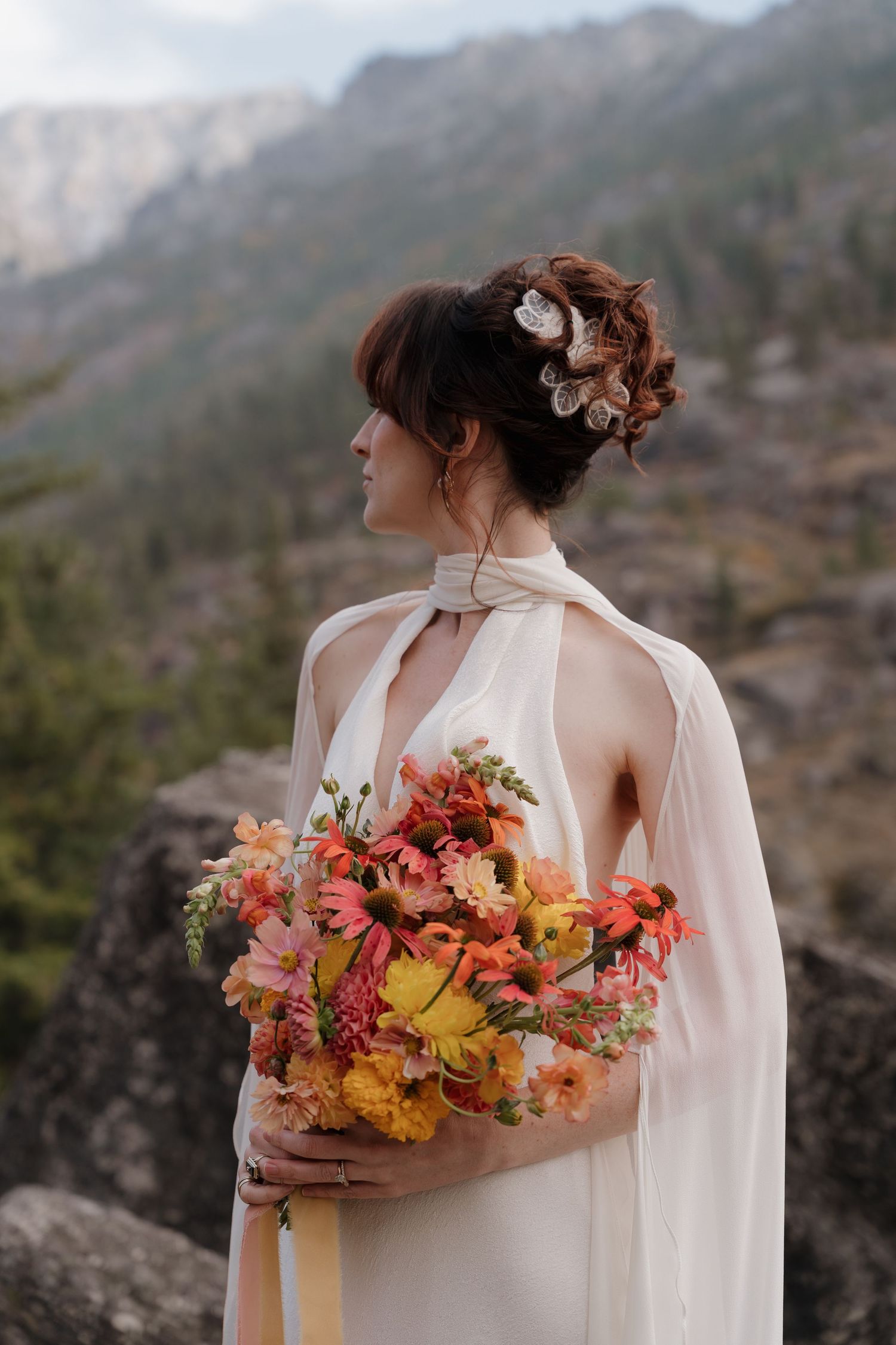 A person in a white flowing dress holds a vibrant autumn-colored bouquet against a mountainous backdrop.